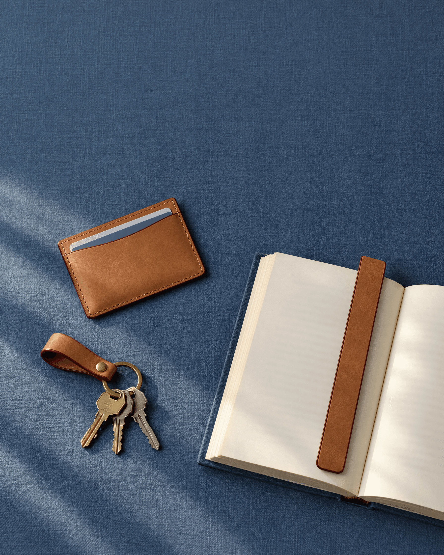 A tan leather card holder, keys on a leather loop and an open book with a leather bookmark, seen from above on slate-blue cloth.